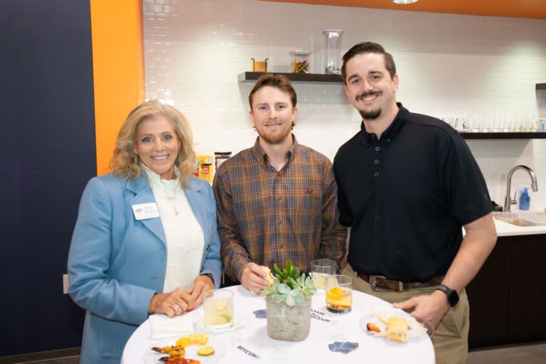 Three people stand around a table with drinks and snacks, smiling at the camera in a room with a kitchenette in the background.