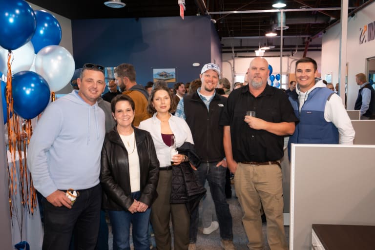 Group of six people posing indoors, with balloons and office decor in the background.