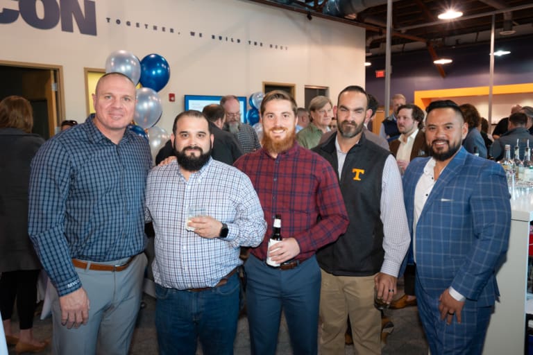 A group of five men posing for a photo at an indoor event with balloons and other attendees in the background.