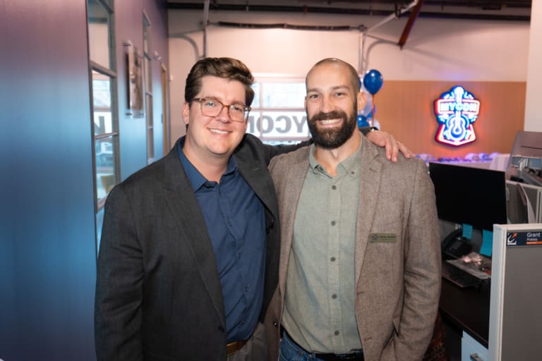 Two men smiling and posing for a photo in a business setting with blue balloons and a neon sign in the background.