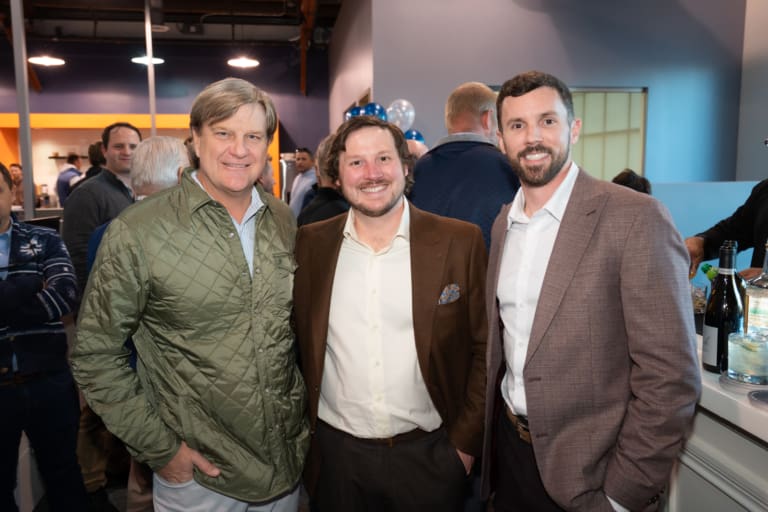 Three men in jackets stand and smile at a gathering. The setting appears to be a casual indoor event.