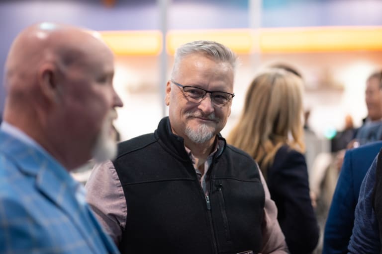 A man with glasses and a goatee smiles while talking to a bald man in a blue jacket at an indoor event.