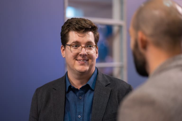 Two people are engaged in a conversation indoors. One person, wearing glasses and a suit jacket, faces the other, who is seen from behind.