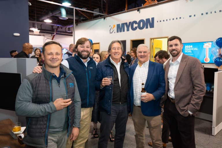 A group of five men pose and smile at an indoor event, holding beverages. A MYCON sign is displayed in the background along with business-themed posters.