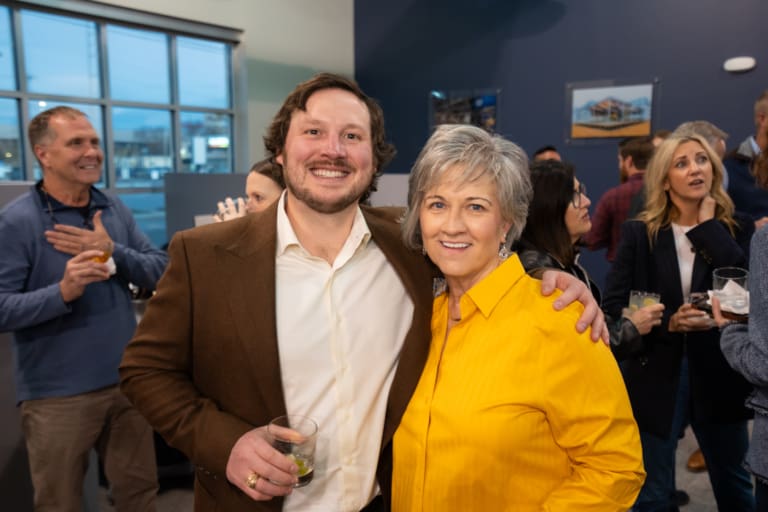 A man and a woman pose smiling at a social event with others in the background, some holding drinks.