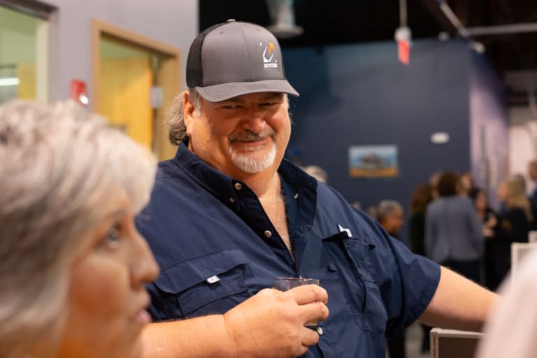 Man wearing a cap and blue shirt holds a drink while smiling at an indoor event. People are mingling in the background.