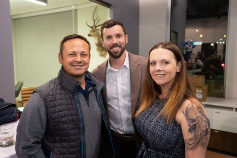 Three people smiling indoors, standing in front of a glass window with a mounted deer head visible on the wall.