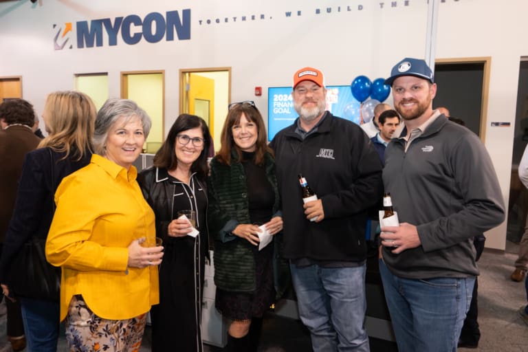 Group of people smiling and holding drinks at an indoor event with a "MYCON" sign in the background.