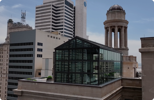 A glass-walled structure on a rooftop with a backdrop of modern and classical buildings in an urban setting under a partly cloudy sky.