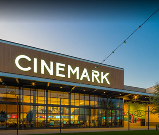 Exterior of a Cinemark theater with large windows and a prominent sign. It is dusk, and lights illuminate the interior and exterior. A popcorn mural is visible on the right side.