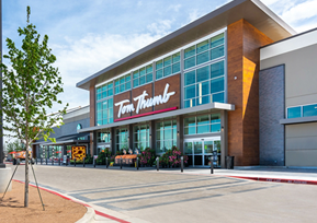 Exterior view of a Tom Thumb grocery store with large windows, a prominent sign, and a few people near the entrance.