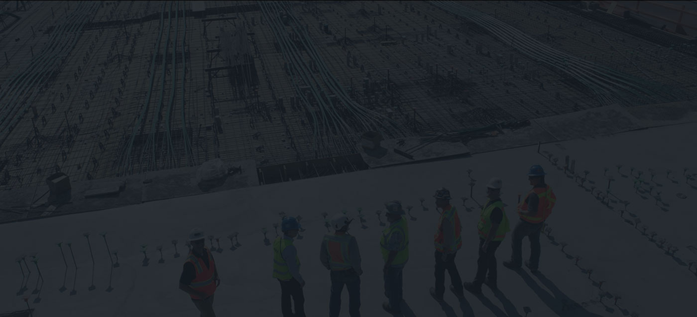 Construction workers in hard hats and safety vests stand on a building site with rebar and concrete.