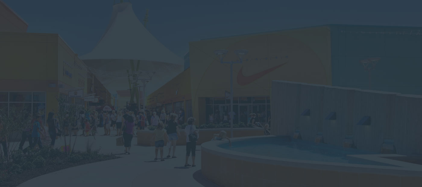 A busy outdoor shopping area with people walking near a circular fountain. Brightly colored store facades are visible under a large canopy.