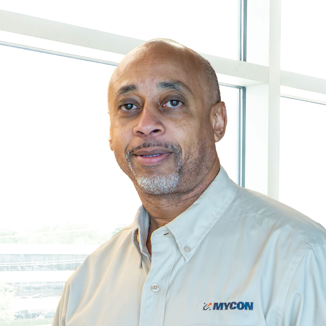 A man wearing a light gray MYCON shirt, stands indoors in front of large windows.
