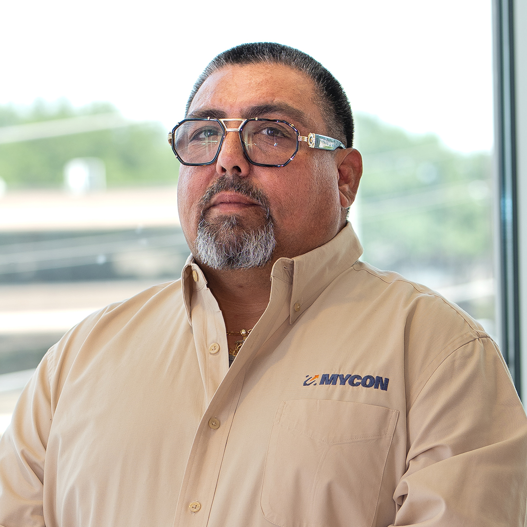 A man wearing glasses and a beige MYCON-branded shirt stands indoors near a window with a blurred outdoor background.