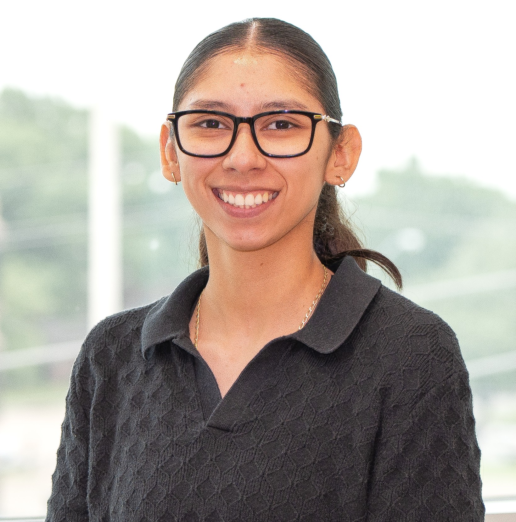 A person with long dark hair tied back, wearing glasses and a black textured shirt, smiles at the camera in front of a bright, blurred outdoor background.