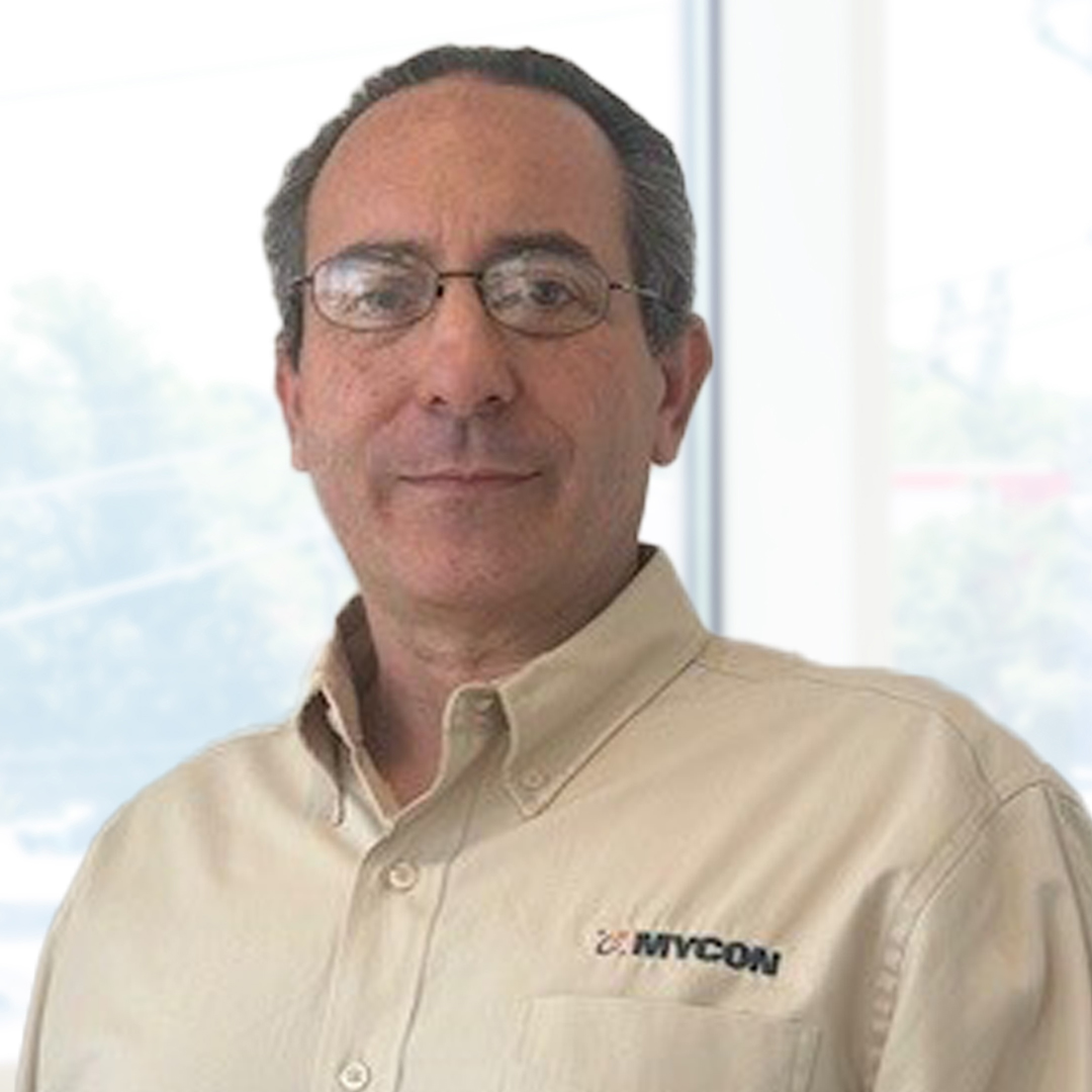 Man with glasses wearing a beige button-up shirt with "MYCON" logo, standing indoors in front of large windows with a blurry outdoor background.