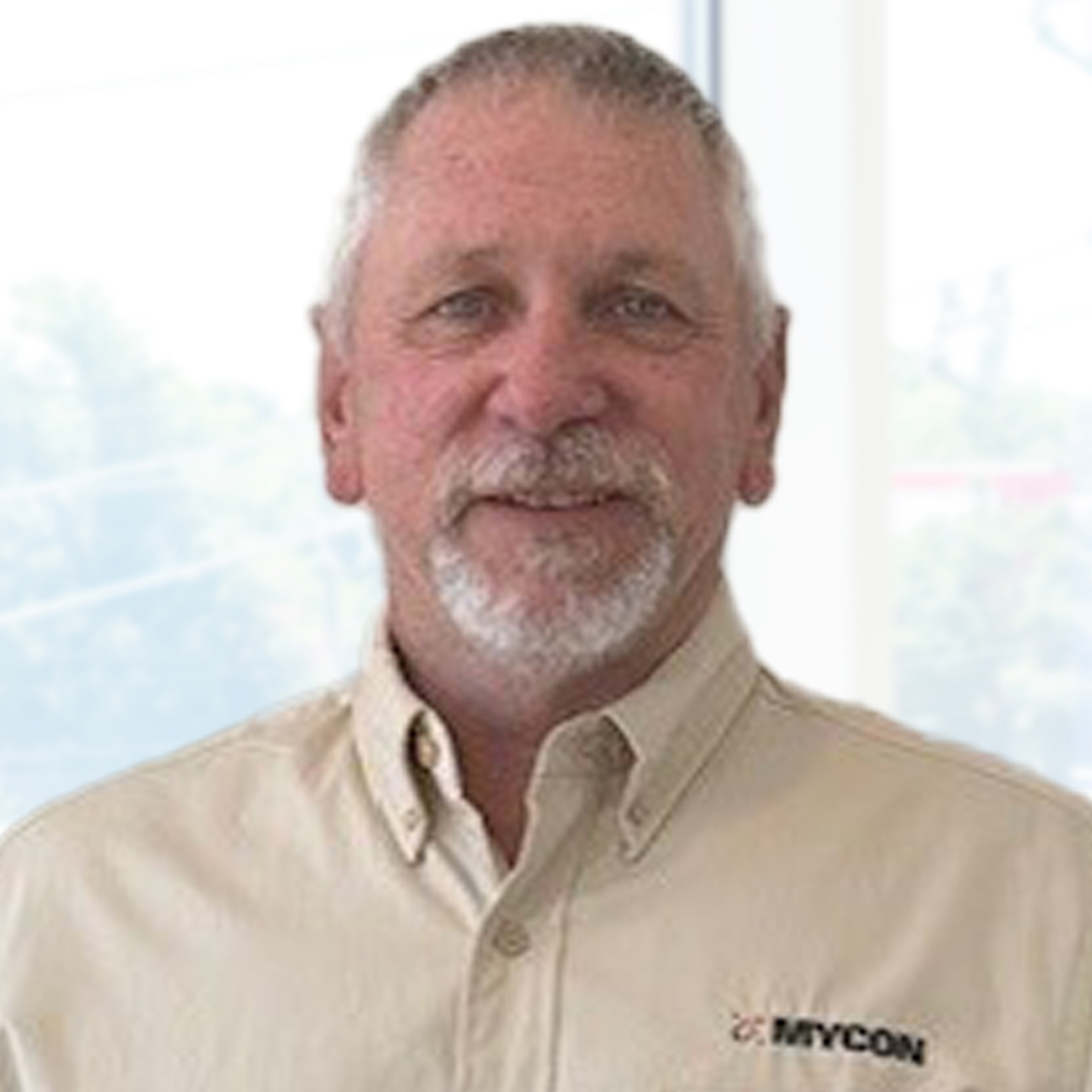 A man with gray hair and a beard wearing a beige button-up shirt with a "MYCON" logo stands indoors in front of a bright window.