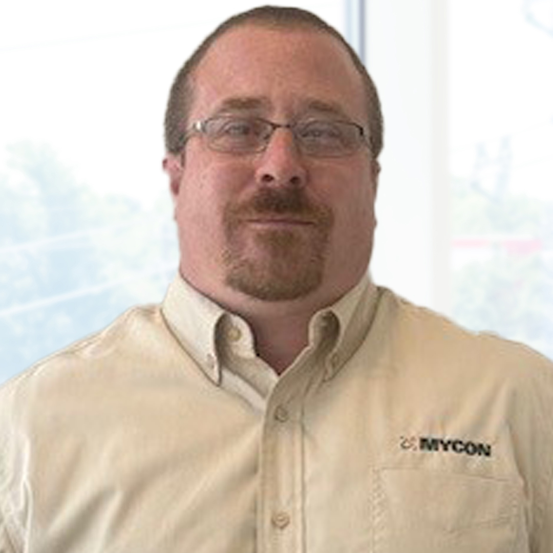 A man with glasses wearing a beige button-up shirt with a "MYCON" logo stands indoors in front of large windows.