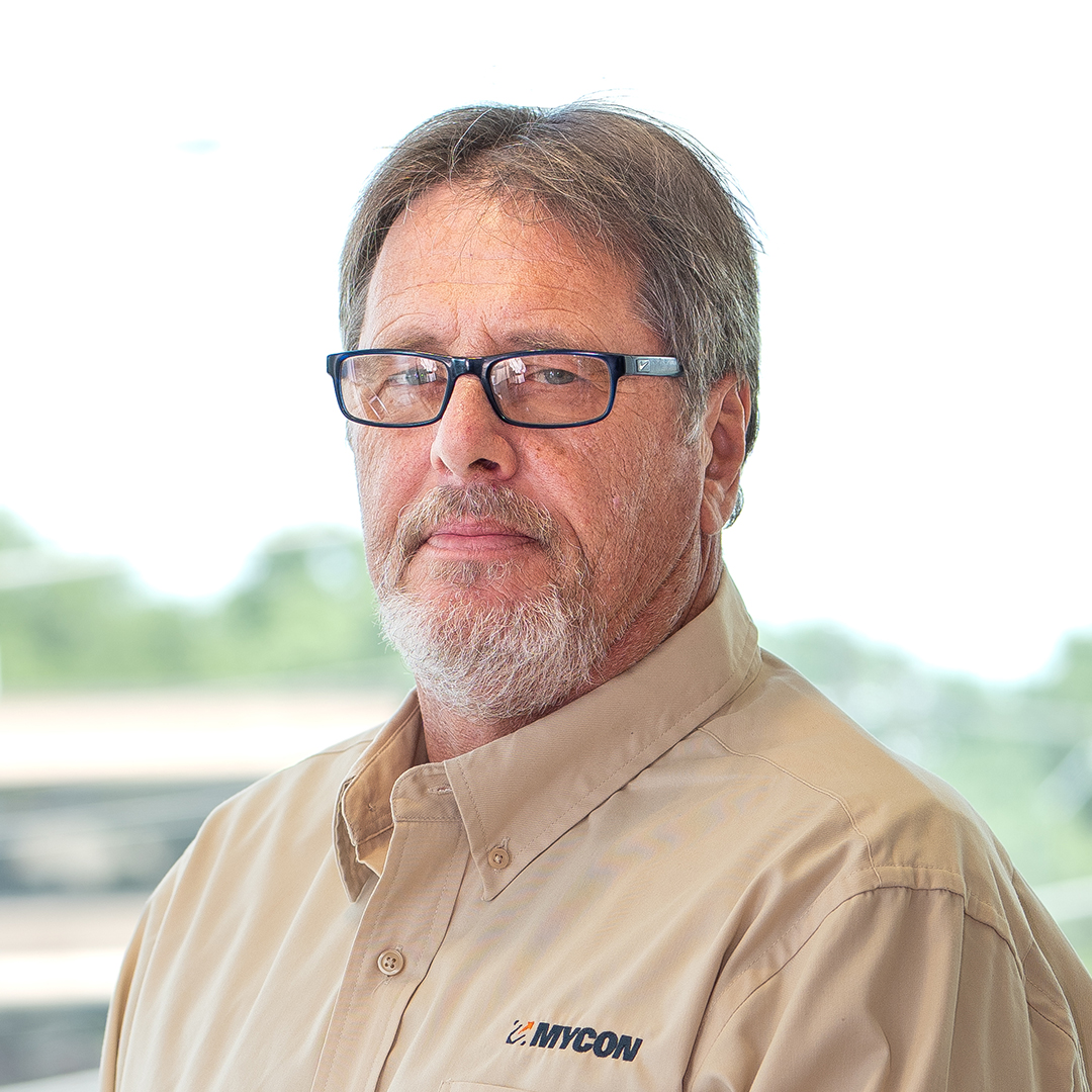 A middle-aged man with glasses, short gray hair, and a beard is wearing a tan MYCON shirt, standing in front of a bright, blurred outdoor background.