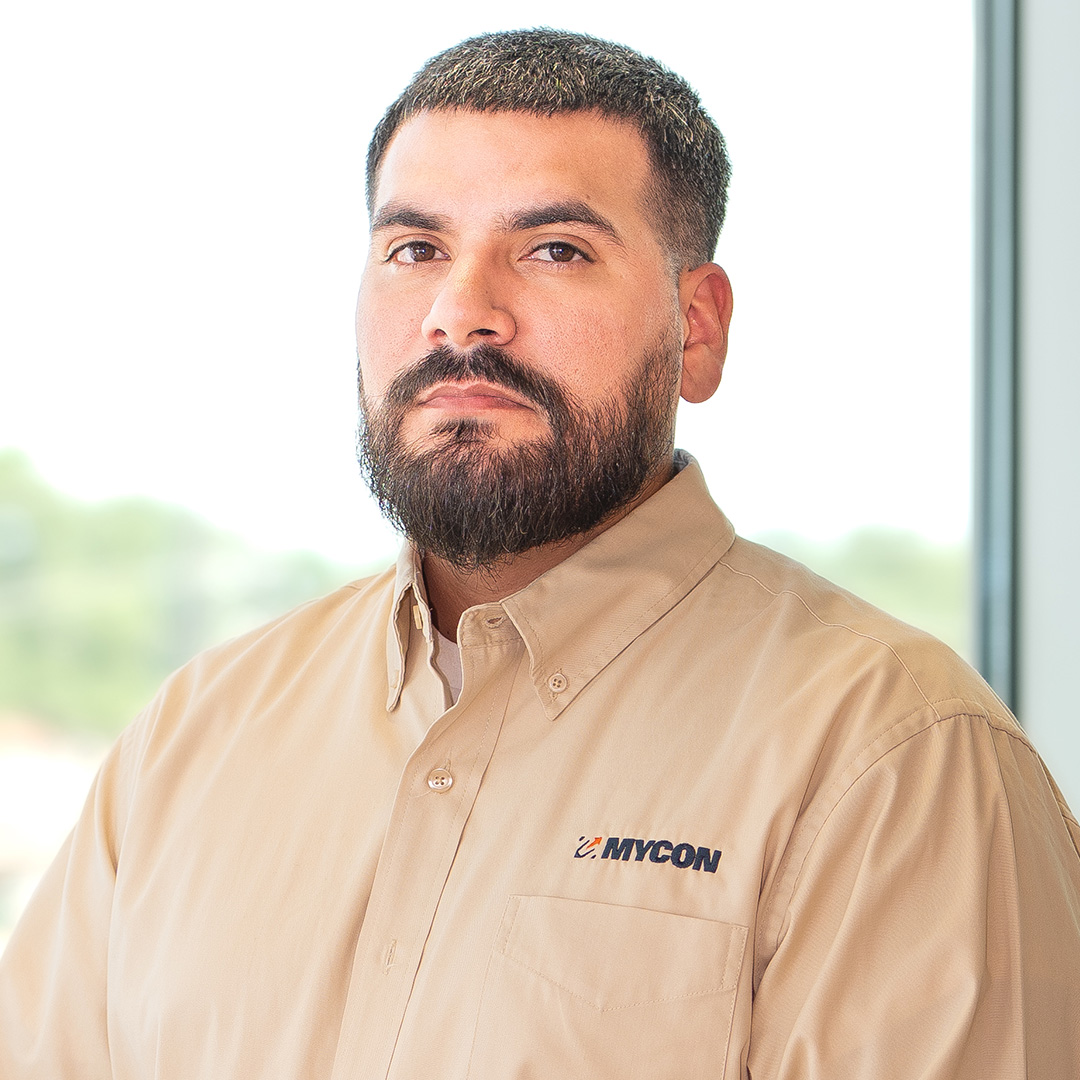 A man with a beard wearing a tan button-up shirt with a MYCON logo stands indoors in front of a window, facing the camera with a neutral expression.