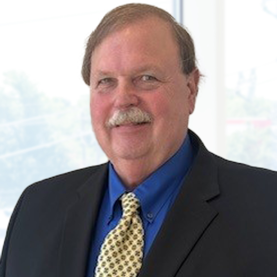 A man with a mustache wearing a dark suit, blue shirt, and patterned tie, standing indoors in front of bright windows.