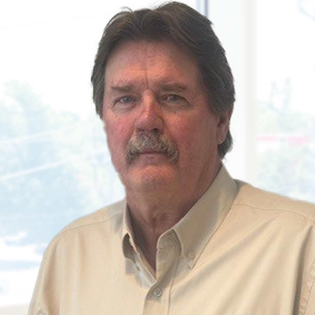 A man with a mustache and medium-length dark hair, wearing a light-colored collared shirt, stands indoors near large windows.