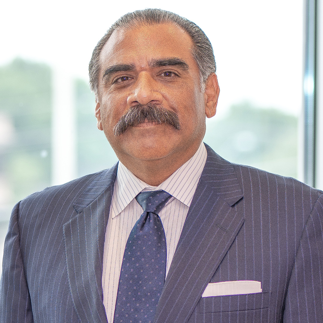A man with a mustache wearing a pinstripe suit, dress shirt, and tie stands in front of a window with a blurred outdoor background.