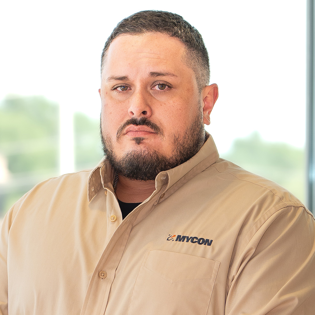 Man with short hair and beard wearing a tan button-up shirt with "MYCON" logo, standing indoors in front of a window with blurred outdoor background.
