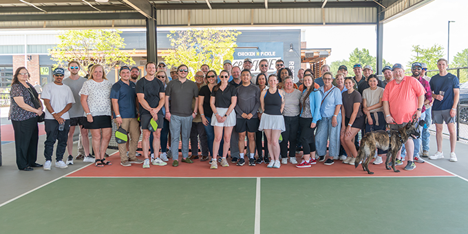 A group of about 35 people, along with one dog, pose together on an outdoor court under a covered area on a sunny day, celebrating their teamwork on recent commercial construction projects.