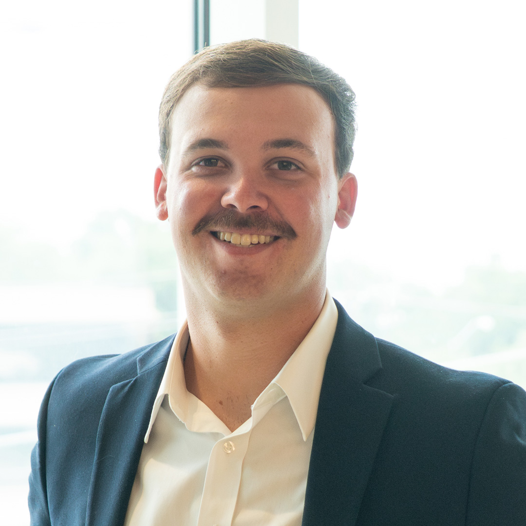 A man with short brown hair and a mustache smiles, wearing a navy blazer and white shirt, standing in front of a bright window.