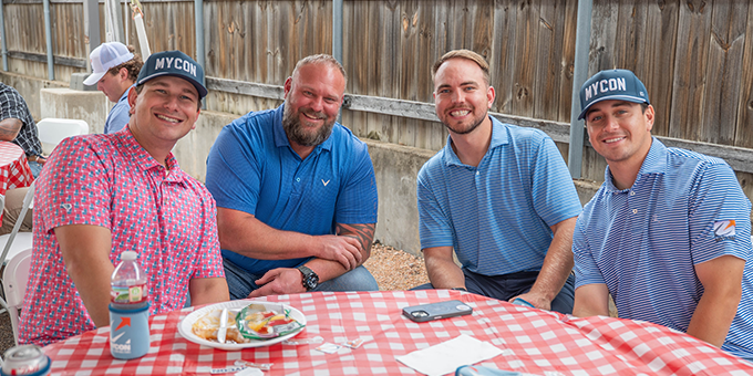 Four men, likely part of a general contractor team, sit at a picnic table with food and drinks, smiling at the camera. Three wear MYCON hats and blue shirts; a wooden fence is in the background.