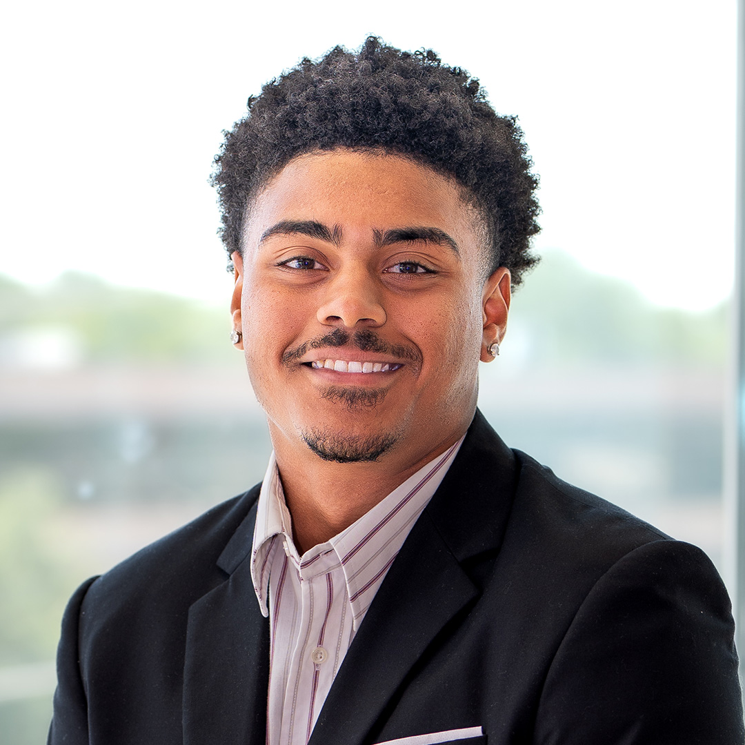 A young man with short curly hair, wearing a black blazer and striped shirt, smiling in front of a blurred outdoor background.