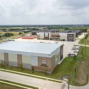 A large storage facility complex, part of the U-Haul of East Denton Construction, features multiple buildings and parking areas next to a multi-lane suburban road under a cloudy sky.