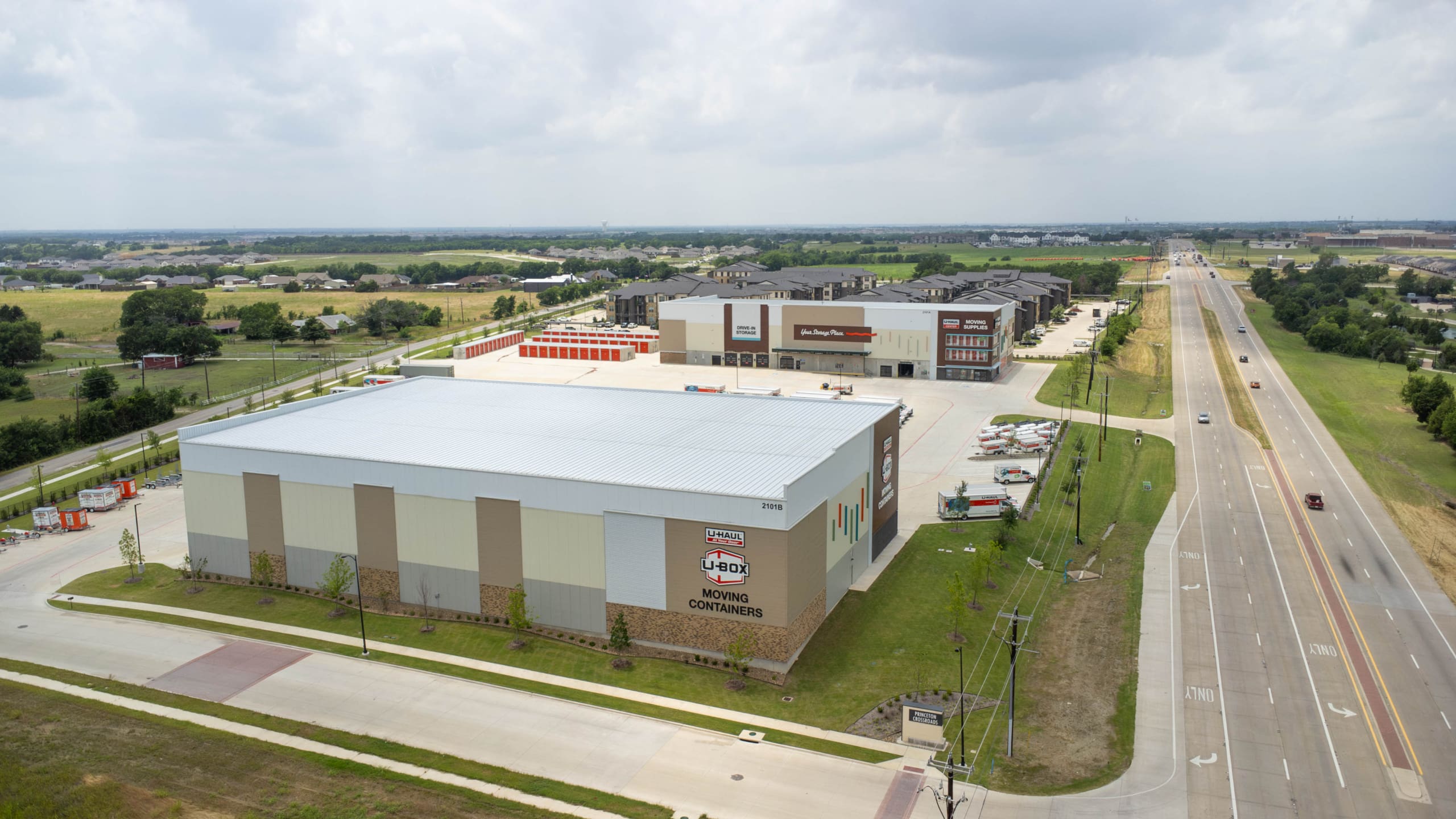 A large storage facility complex, part of the U-Haul of East Denton Construction, features multiple buildings and parking areas next to a multi-lane suburban road under a cloudy sky.