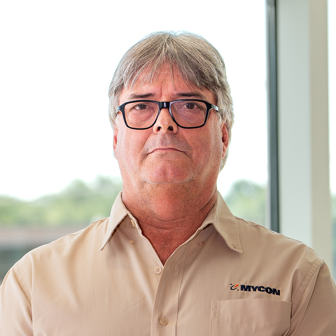 A middle-aged man with glasses and gray hair wearing a beige MYCON shirt stands indoors near a window, facing the camera with a neutral expression.