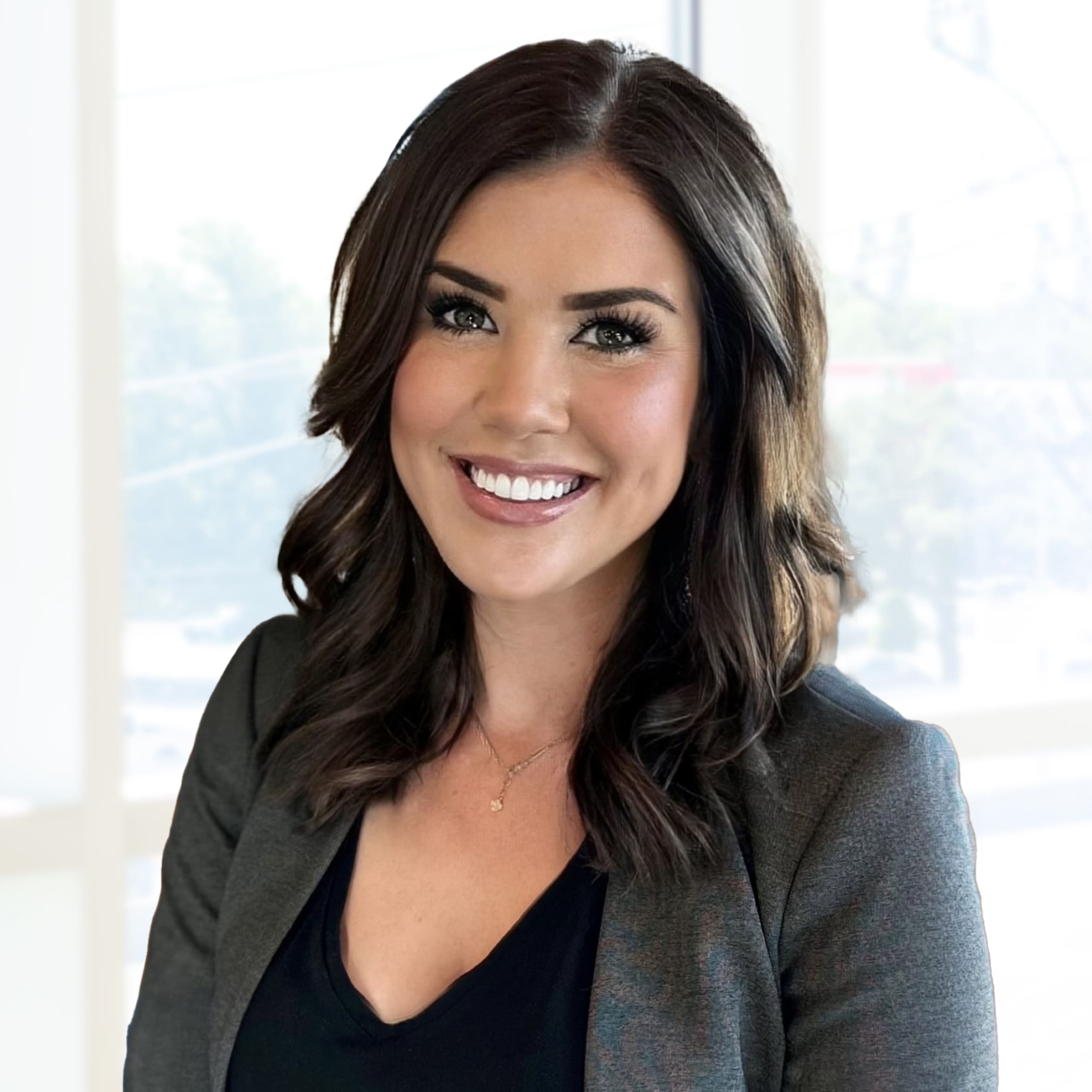 A woman with dark hair in loose waves, wearing a gray blazer over a black top, smiles at the camera in a bright office setting.