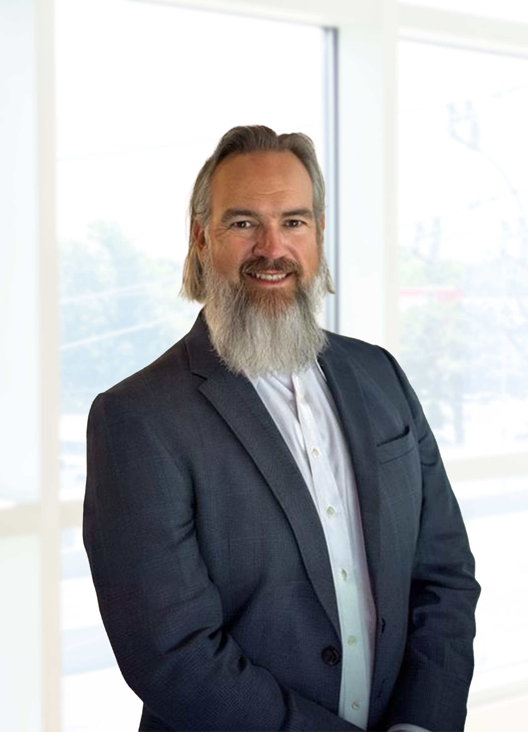 A man with a long gray beard and shoulder-length hair, wearing a dark suit and white shirt, stands in front of large windows in a bright office setting.