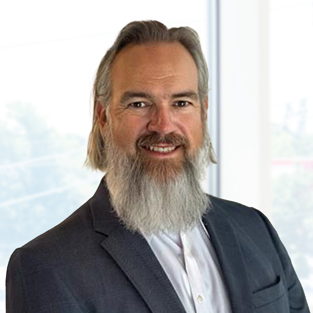 A man with long gray hair and a full gray beard is wearing a dark suit jacket and white shirt, standing in front of a bright window.