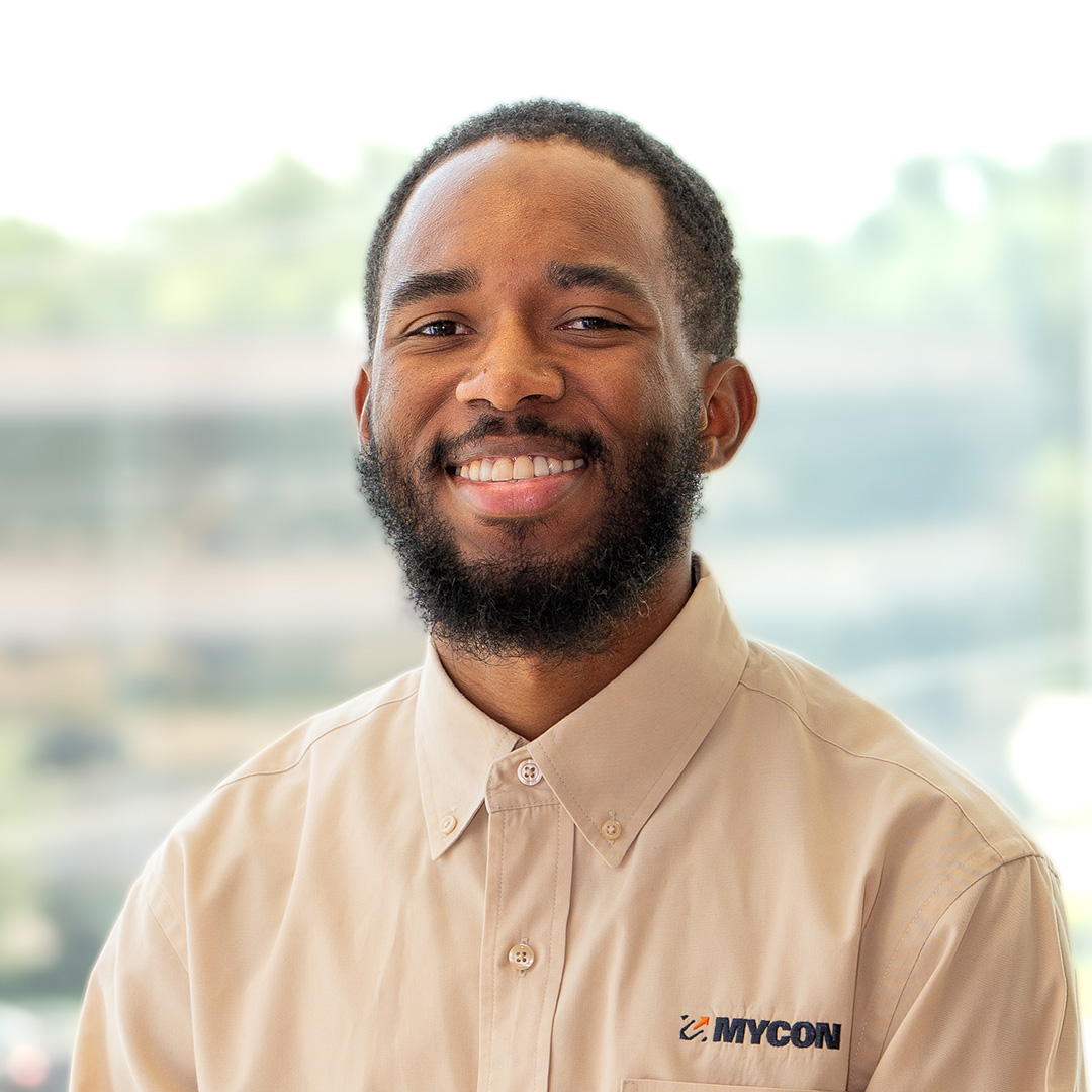 A man with a beard smiles at the camera, wearing a tan button-up shirt with a "MYCON" logo, against a blurred outdoor background.