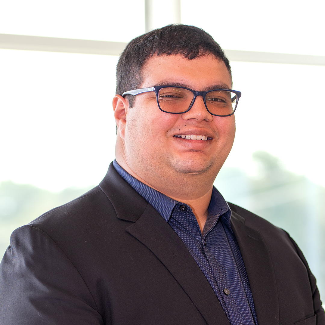 A man wearing glasses, a dark blazer, and a blue shirt smiles while standing indoors in front of a bright window.