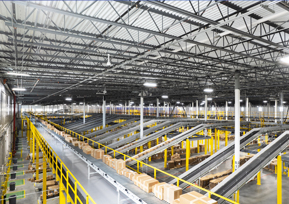 Large warehouse interior with conveyor belts transporting cardboard boxes, surrounded by yellow safety railings and high industrial ceilings—showcasing our retail construction expertise.