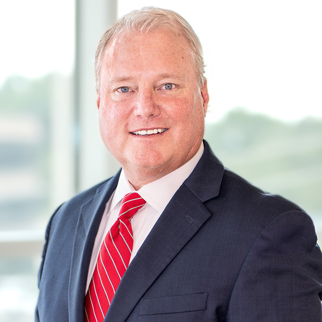 A man in a navy suit, white shirt, and red striped tie stands indoors, smiling at the camera with a blurred office window background.