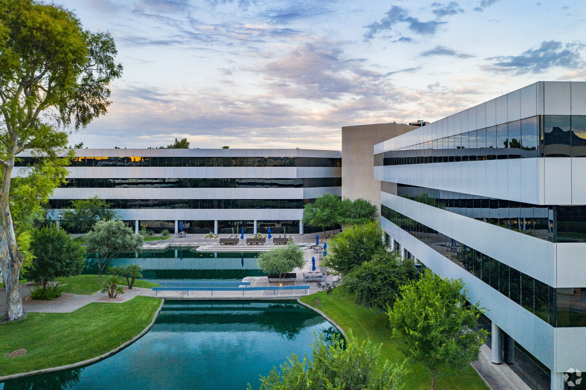 Modern office buildings with mirrored windows surround a landscaped courtyard featuring a pond and walking paths, designed with input from contractor consultation, under a partly cloudy sky.