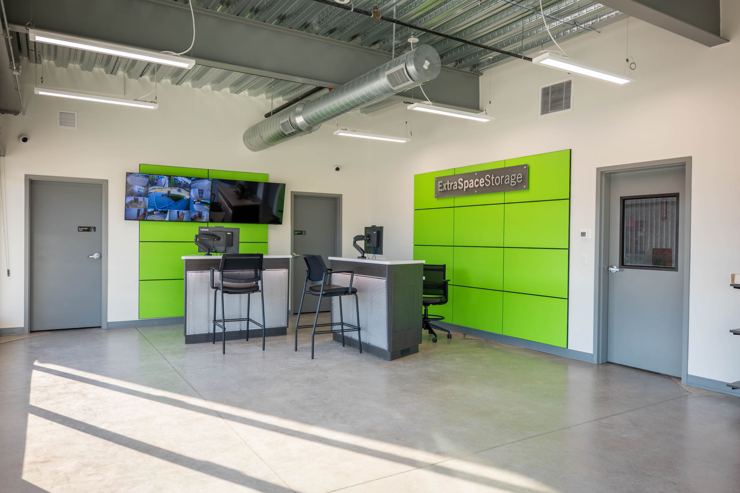 A modern storage facility office features bright green accent walls, two desks with computers, comfortable chairs, and a wall-mounted TV displaying images.