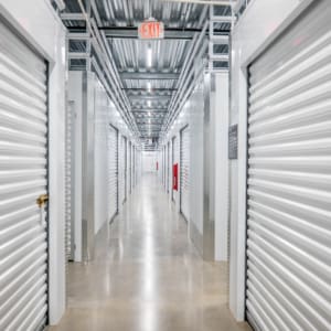 A brightly lit hallway inside self-storage facility, featuring rows of closed white roll-up doors on both sides and a polished concrete floor.
