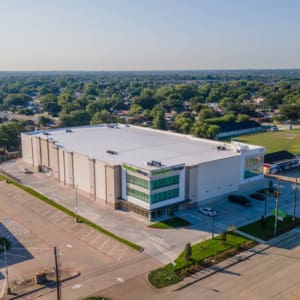 A large, white commercial building with a flat roof and green-tinted windows—likely an Extra Space Storage in Allen, TX—is surrounded by an empty parking lot and suburban houses.