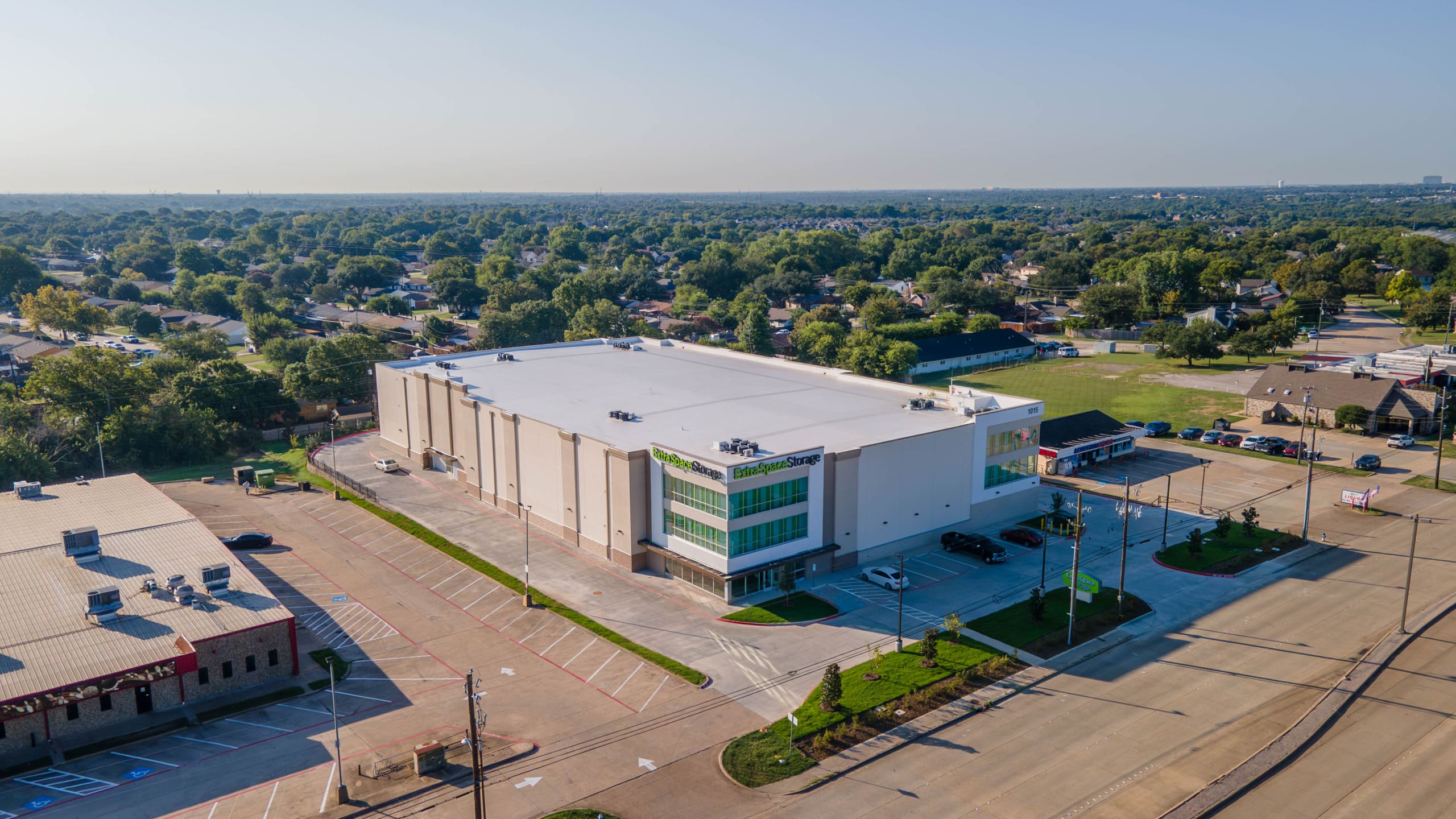 A large, white commercial building with a flat roof and green-tinted windows—likely an Extra Space Storage in Allen, TX—is surrounded by an empty parking lot and suburban houses.