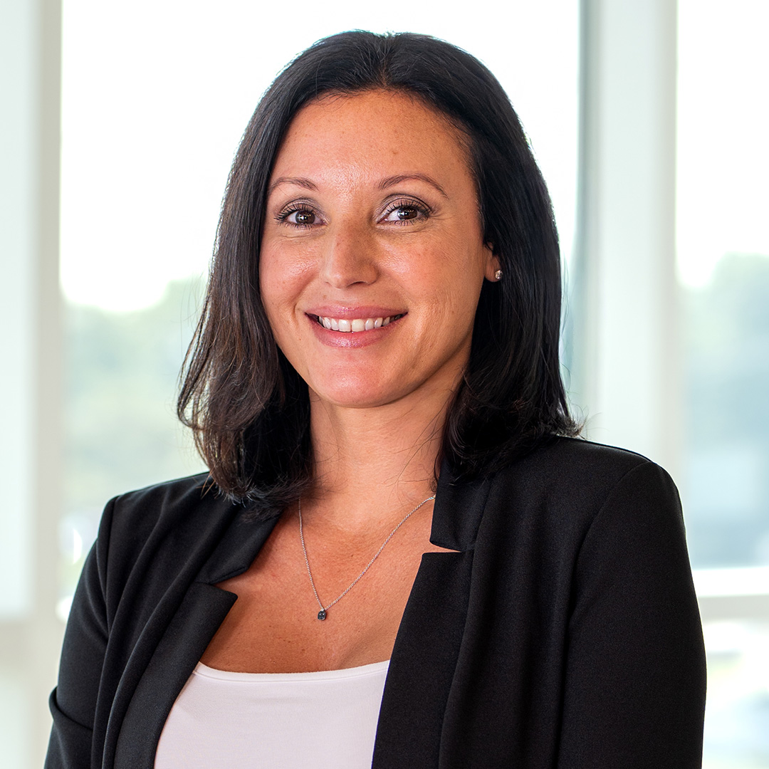 Woman with shoulder-length dark hair wearing a black blazer and white top, smiling while standing indoors in front of large windows.
