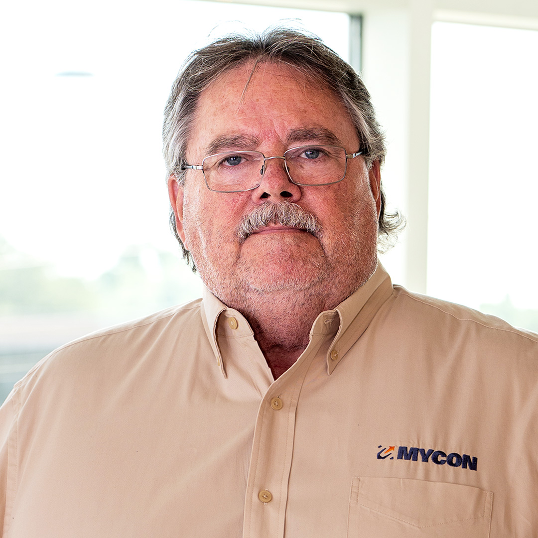 Man with glasses and mustache wearing a beige MYCON shirt stands indoors in front of bright windows.